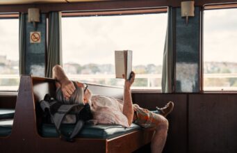 Wie Slow Travel das Urlaubsverhalten prägt Slow Travel: Ein Mann liegt auf einer Sitzbank im Zug, hat ein Buch in der Hand und blickt durch das große Fenster in die Landschaft