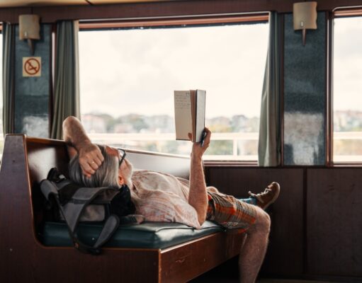 Wie Slow Travel das Urlaubsverhalten prägt Slow Travel: Ein Mann liegt auf einer Sitzbank im Zug, hat ein Buch in der Hand und blickt durch das große Fenster in die Landschaft