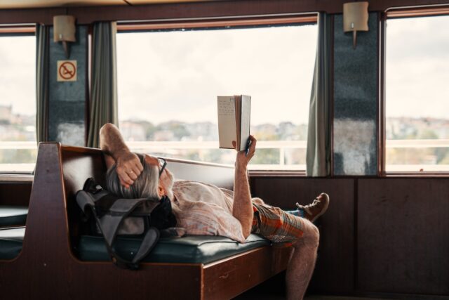Slow Travel: Ein Mann liegt auf einer Sitzbank im Zug, hat ein Buch in der Hand und blickt durch das große Fenster in die Landschaft